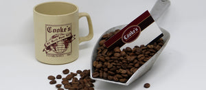   A beige mug, printed with the Cooke's Old World Shop logo, beside a silver scoop of dark brown coffee beans. In the coffee beans sits a Cooke's business card. All on a white background, with dark brown coffee beans scattered. 