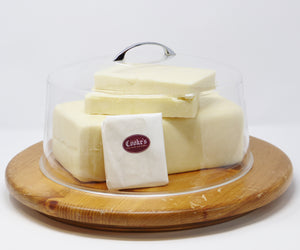 Cooke's white cheddar in three large blocks, with one small piece wrapped in white cheese paper and stickered with the Cooke's burgundy logo, all under a clear dome with metal handle and sitting on a round wooden board.